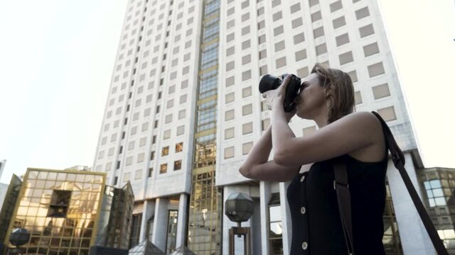 Female photographer with professional camera. Action. Young woman holding camera in her hands and taking pictures on street walk on the background of a building with many windows and golden glass