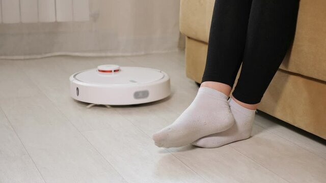 Young woman wearing white socks sits on sofa and lets round automatic robot vacuum cleaner pass feet on wooden floor in living room close view