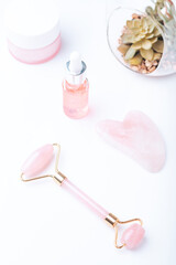 Pink hyaluronic acid serum, rose quartz beauty roller, gua sha and flowers on pink table top. Anti aging cosmetics and tools, copy space, top view