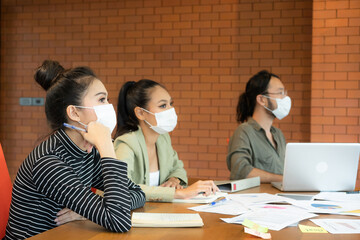 Group of young Asian business people in protective face mask for prevent coronavirus infection sitting at desk with notepad, pie chart paper, computer notebook and paying attention during meeting.
