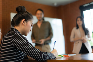 Asian businesswoman in long sleeve striped shirt sit at desk and taking notes during her asian colleagues giving presentation about the new business plan on whiteboard in the office meeting room.