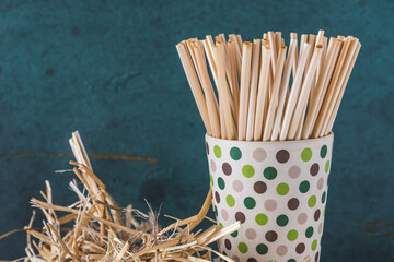 Natural straw drinking straws in a bamboo beaker and a bundle of straw on an old wooden beam, green background. Concept of sustainability, no plastic. Closeup with copy space.