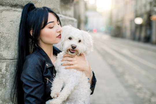 Portrait of young pretty Asian woman with black ponytail hairstyle and bright makeup, smiling with closed eyes, holding her cute white dog, standing near old building in the city. Close up portrait