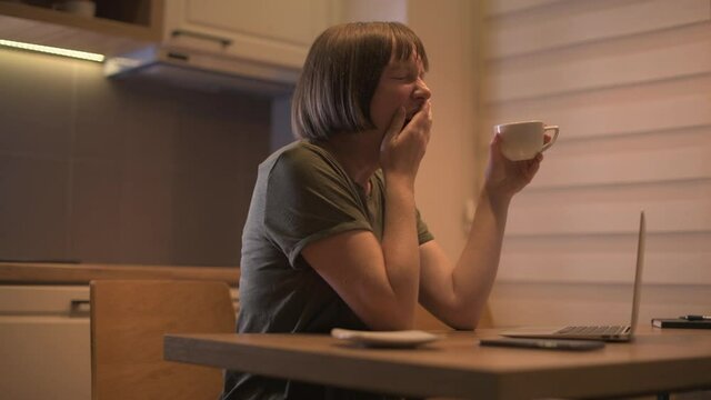 Tired exhausted female freelancer working from home, drinking coffee and yawning