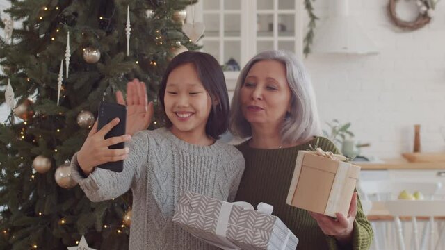 Joyous senior grandma and cheerful little girl of Asian ethnicity holding gift boxes, smiling and waving at smartphone camera while taking selfie or video calling beside Christmas tree at home