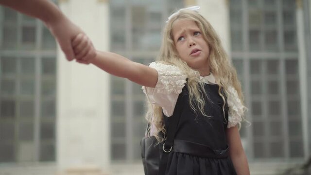 Portrait of cute stubborn schoolgirl unwilling to go to school in the morning. Unrecognizable brother pulling little sister's hand. Schoolchildren lifestyle and education.