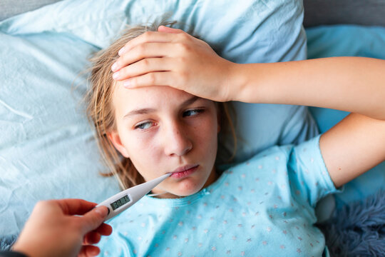 Sick teenage little girl with high fever and headache laying in bed and holding thermometer in her mouth