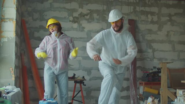 Young funny female and male worker in white protective suits dancing at construction site. Positive house renovation process. People in protective hardhats. "Thermoset powder blending" sign