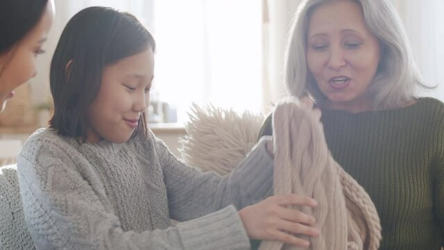 Happy senior woman taking warm handmade scarf form gift box, saying thanks and embracing cute Asian granddaughter and cheerful daughter on sofa in living room while celebrating Christmas together