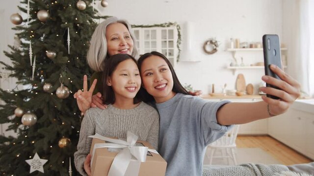 Cheerful Asian woman, her little daughter and grandma smiling and posing together for smartphone camera while taking a selfie against Christmas tree at home