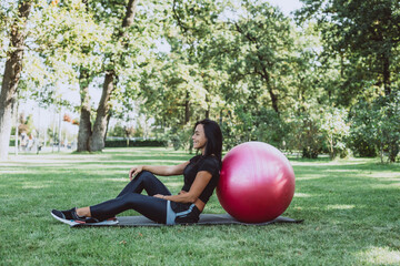 Young happy fit tanned brunette goes in for sports with a big fitness ball, dressed in active black clothes, does exercises, works out, posing in the park, walks outdoors. Soft selective focus.