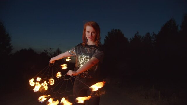 Elegant lovely redhead female fire juggler performing impressive stunt with two burning metal fans outdoors at night, Graceful dancer with fans lit with flames juggling during fire show performance.