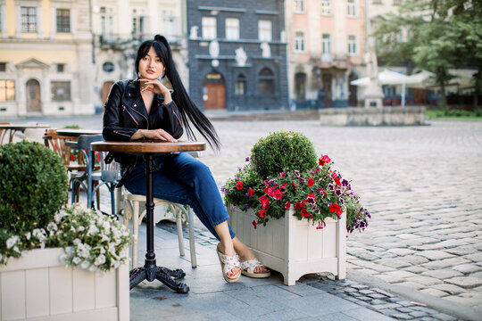 Happy Asian brunette woman at outdoors cafe, sitting at the table. Young stylish woman in jeans and leather jacket, resting outdoors at the cafe in old European city