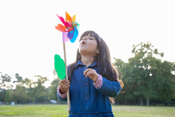 Adorable black haired girl holding pinwheel and blowing on toy, playing in park. Medium shot, front view. Children outdoor activity concept