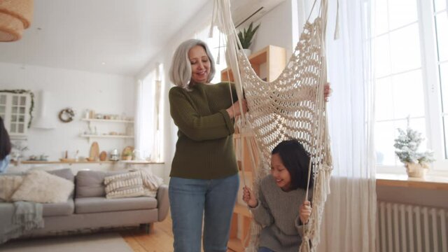 Little Asian girl smiling and swinging on indoor macrame swing with assistance of cheerful grandmother while mother decorating Christmas tree in background