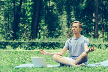Obraz premium Young attractive man in sport clothes is meditating in the lotus position with a pacified face in the park against the background of green grass and trees.
