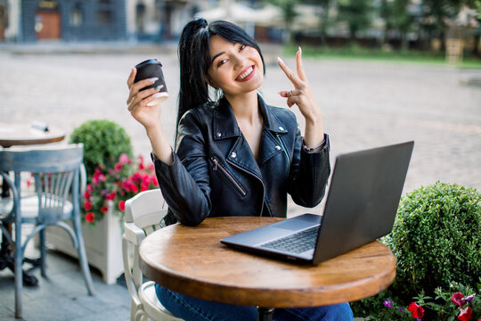 Beautiful smiling cute asian young businesswoman, dressed in black leather jacket, with ponytail hair, sitting at the table in outdoor cafe, using laptop, drinking coffee, and showing victory sign