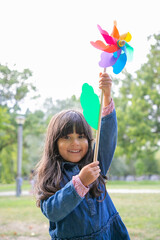 Happy black haired girl playing in park, holding pinwheel, looking at camera and smiling. Front view, vertical shot. Children outdoor activity concept
