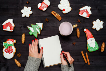 Woman with red manicure writing New year resolutions in blank notebook, copy space. Christmas background with gingerbread cookies, cinnamon and pine cones on dark wooden desk. Top view, flat lay