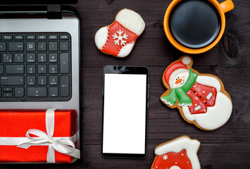 Laptop computer, smartphone with blank screen, Christmas present, cup of coffee and  gingerbread cookies on dark wood background, copy space. Top view, flat lay.