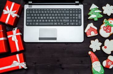 Fototapeta na wymiar Open laptop computer, Christmas presents and gingerbread cookies on dark wood background, copy space. Top view, flat lay. Santa, snowflake, mitten, house, stocking, snowman, fir tree