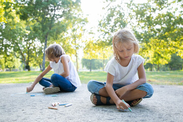 Two focused cute girls sitting and drawing with colorful pieces of chalks. Front view. Childhood and creativity concept