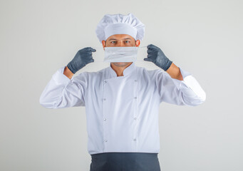 Male chef wearing medical mask in uniform, apron and hat and looking careful. front view.