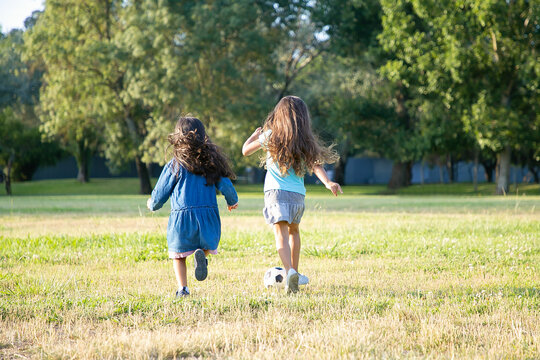 Active black haired girls running for soccer ball on grass in city park. Full length, back view. Childhood and outdoor activity concept