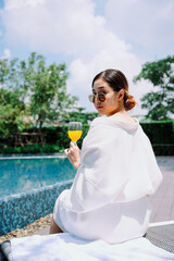 Asian woman traveler in white bathrobe enjoy drinking orange juice at poolside outdoors.