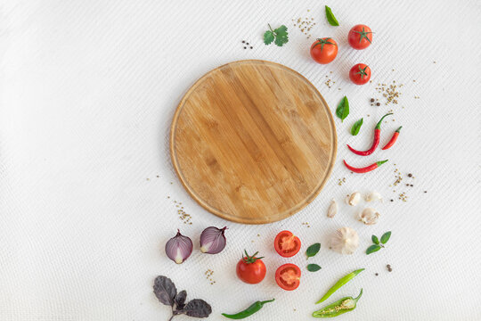 Empty round wooden cutting board on white textured background with spices, onion, garlic, chilli pepper, cherry tomatoes, basilic and greens with copy space. Top view.
