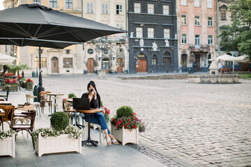 Obraz premium Young attractive brunette woman using laptop at outdoor city cafe, sitting at the table and drinking coffee. Old vintage buildings of European city on the background