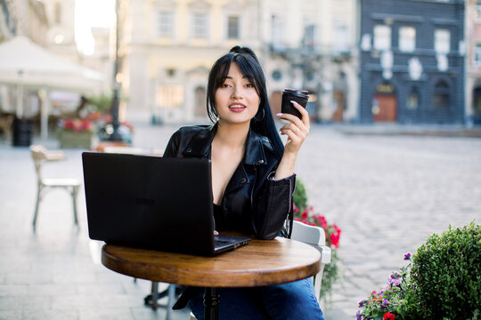 Freelance work concept. Young attractive Asian woman wearing black leather jacket, working on a laptop and drinking coffee, sitting at the table in outdoor city cafe