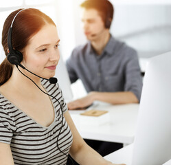 Casual dressed young woman using headset and computer while talking with customers online. Group of...