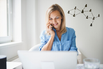 beautiful young businesswoman in blue shirt is sitting behind her laptop and is using her smartphone in her home office