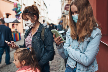 Young women using smartphones social media sending texts watching photos and videos talking remotely with friends standing in a crowded street downtown. Woman is wearing the face masks to avoid virus