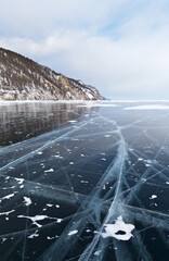 Beautiful winter landscape of frozen Baikal Lake. Snow-covered hills on the shore and blue transparent ice with cracks. Ice travel. Natural cold background
