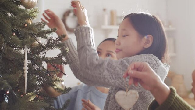 Cute little Asian girl decorating Christmas tree with help of happy young mother and grandmother while preparing home for holiday