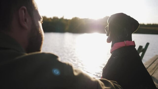 young guy sitting with dog on the pier and admiring the beauty of the lake