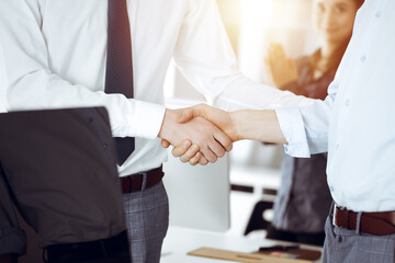 Two businessmen shaking hands in sunny office, close-up. Happy and excited business woman stands with raising hands at the background. Business people concept