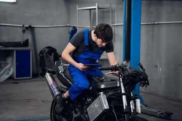 A repairer in work clothes is sitting on a bike. He uses a tablet