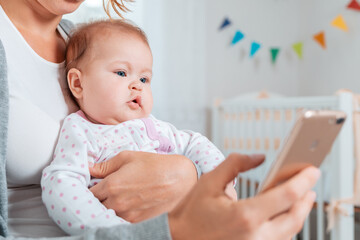Portrait of a baby sitting in his mother's arms and looking at the smartphone that she uses. Concept of teaching and entertaining children using the Internet