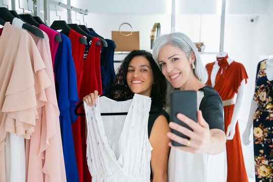 Cheerful cute female friends enjoying shopping in boutique together, holding dress, having fun and taking selfie on mobile phone. Consumerism or shopping concept