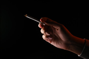 Woman hand holding cigarette in a smoke against black background. Smoking cannabis joint. Medical use.	