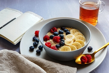 Side view of a healthy breakfast with oatmeal in a bowl, banana slices, raspberry, blueberry, a cup of tea and a notebook with a pen on light grey tabletop
