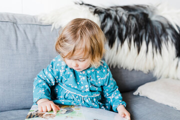 Toddler girl sitting on sofa and reading picture book