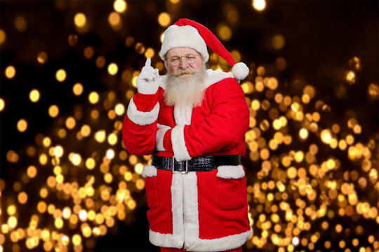 Santa Clause raised index finger. Bearded concentrated Santa Claus pointing with index finger upwards standing on New Year lights background.