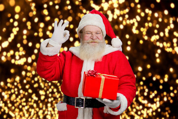 Authentic Santa Claus with okey gesture. Happy Santa Claus holding red gift box and showing ok sign on New Year lights background.