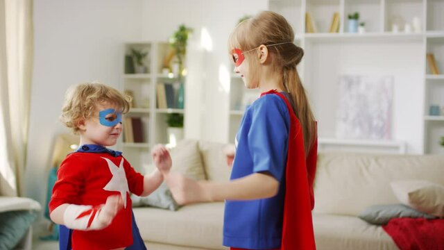 Medium closeup of young brother and sister in superhero costumes with eye masks and wristbands standing in front of each other playing patty-cake with hands