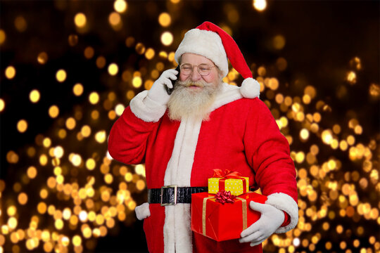 Santa Claus having Christmas call. Happy Santa Claus holding present boxes and talking on mobile phone on festive lights background.