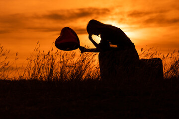 Silhouette of a sad woman sitting on suitcase with heart shaped balloon  at the sunset.
